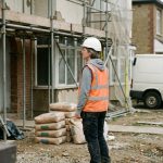 Cheffe de chantier en gilet et casque devant une maison en rénovation, échafaudage et sacs de ciment au premier plan